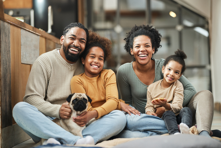 A happy family of four sits together on the floor in a cozy home, smiling warmly at the camera