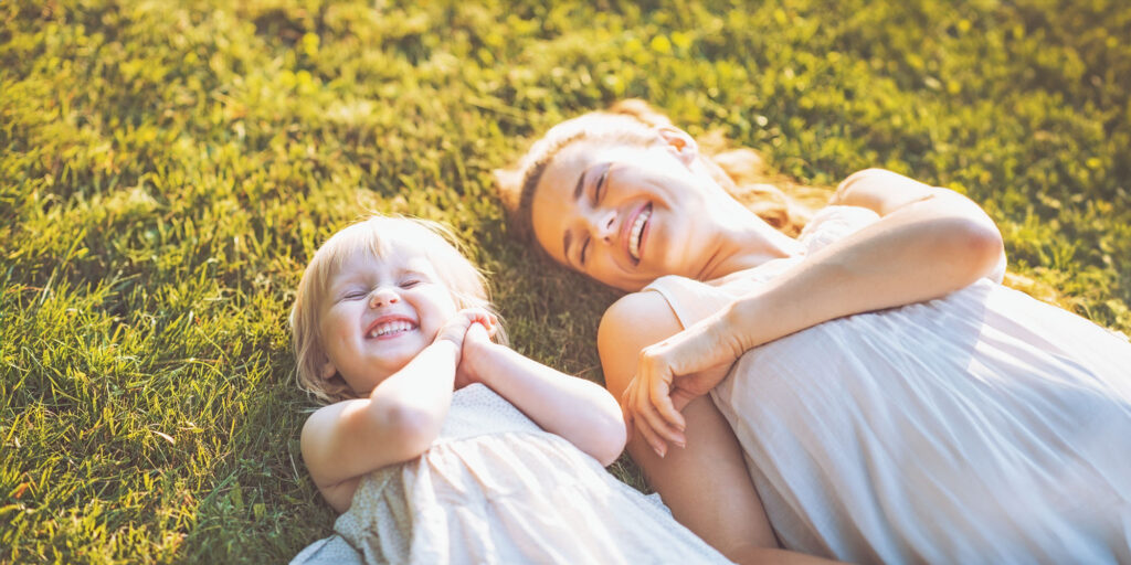 Heartwarming scene of a joyful mother and her young daughter lying on their backs in a sunlit grassy field, both smiling brightly at each other, capturing a tender moment of shared happiness.